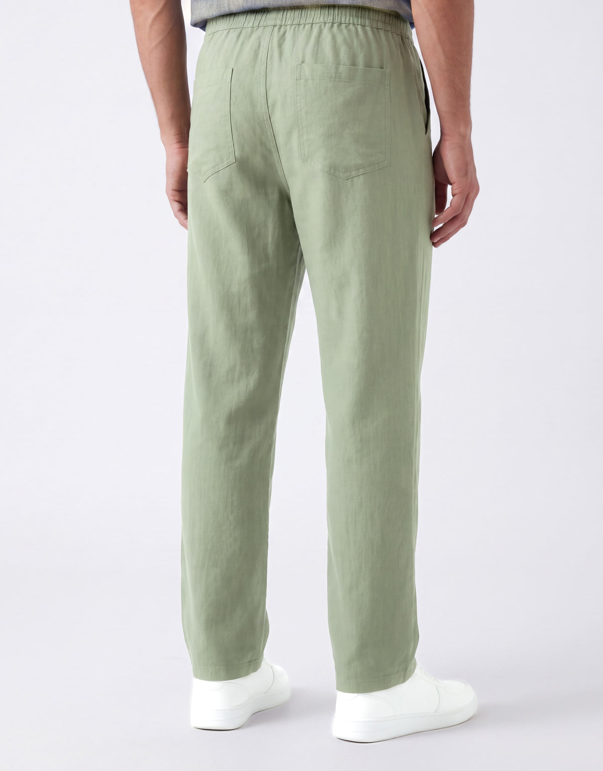 A person shown from the back wearing light sage green linen trousers and white sneakers against a clean white studio background.