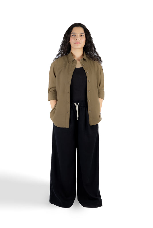 A woman stands against a white studio background wearing a brown button-up shirt over a black top and black wide-leg pants.