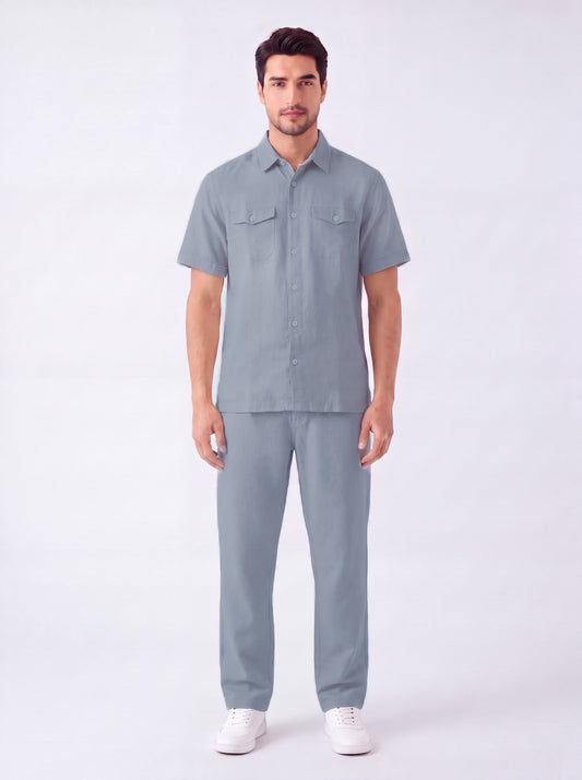 A man modeling a matching light blue-gray short-sleeve button-up shirt and trousers set, paired with white sneakers, against a plain white studio background.