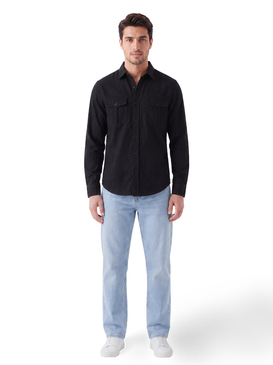 A man modeling a black long-sleeve button-up shirt with two chest pockets, paired with light blue jeans and white sneakers, against a plain white studio background.