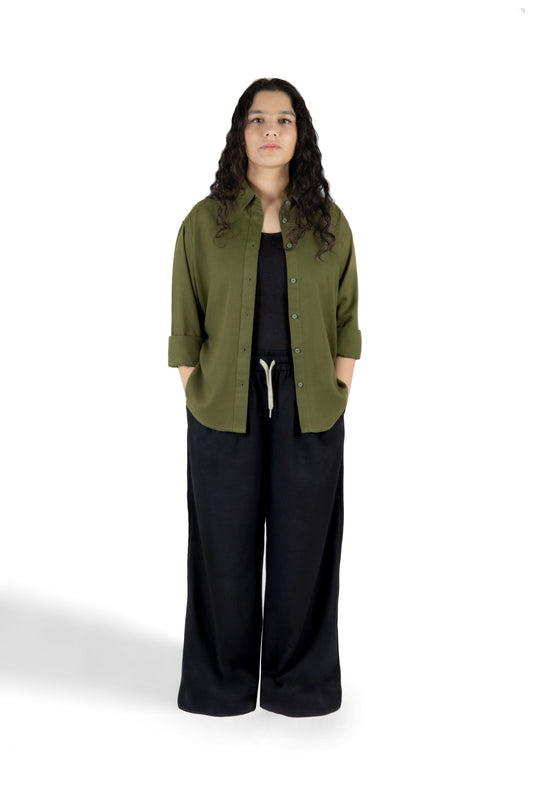 A woman stands against a white studio background wearing an olive green button-up shirt with three-quarter sleeves over a black top and black wide-leg pants.