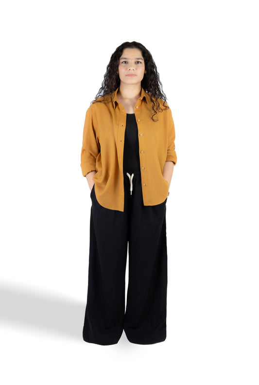 A woman modeling a mustard yellow button-up shirt over a black top and black wide-leg pants, standing against a plain white studio background.