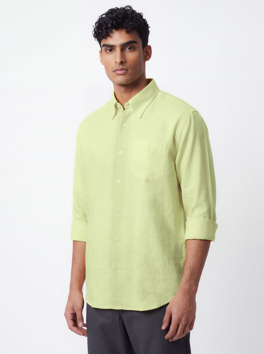 A man wearing a light yellow-green long-sleeve button-down shirt with rolled-up sleeves and dark gray trousers, standing against a plain white studio background.