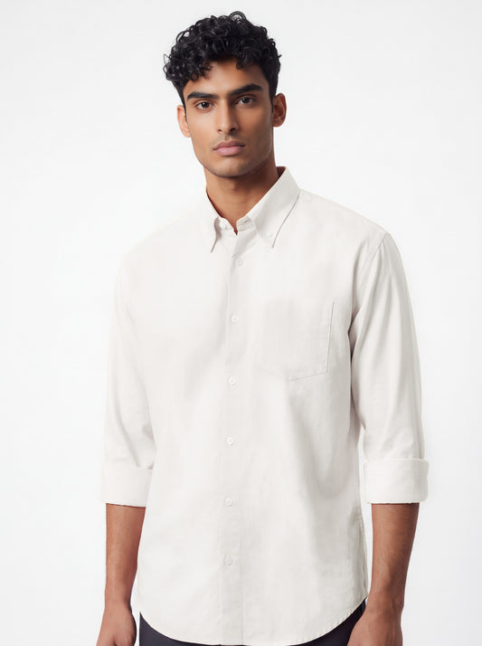 A man wearing a light beige long-sleeve button-down shirt and dark gray trousers, standing against a plain light gray studio background.