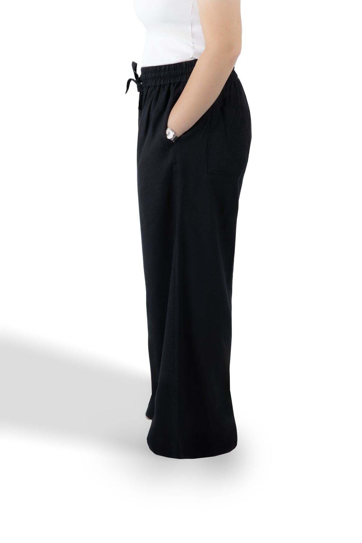 A side profile view of a person wearing black wide-leg lounge pants with a drawstring waist and side pockets, paired with a white top, against a white studio background.