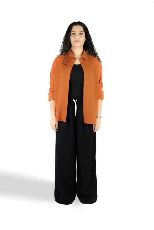 A woman stands against a white studio background wearing a rust-orange button-up shirt over a black top and black wide-leg pants.