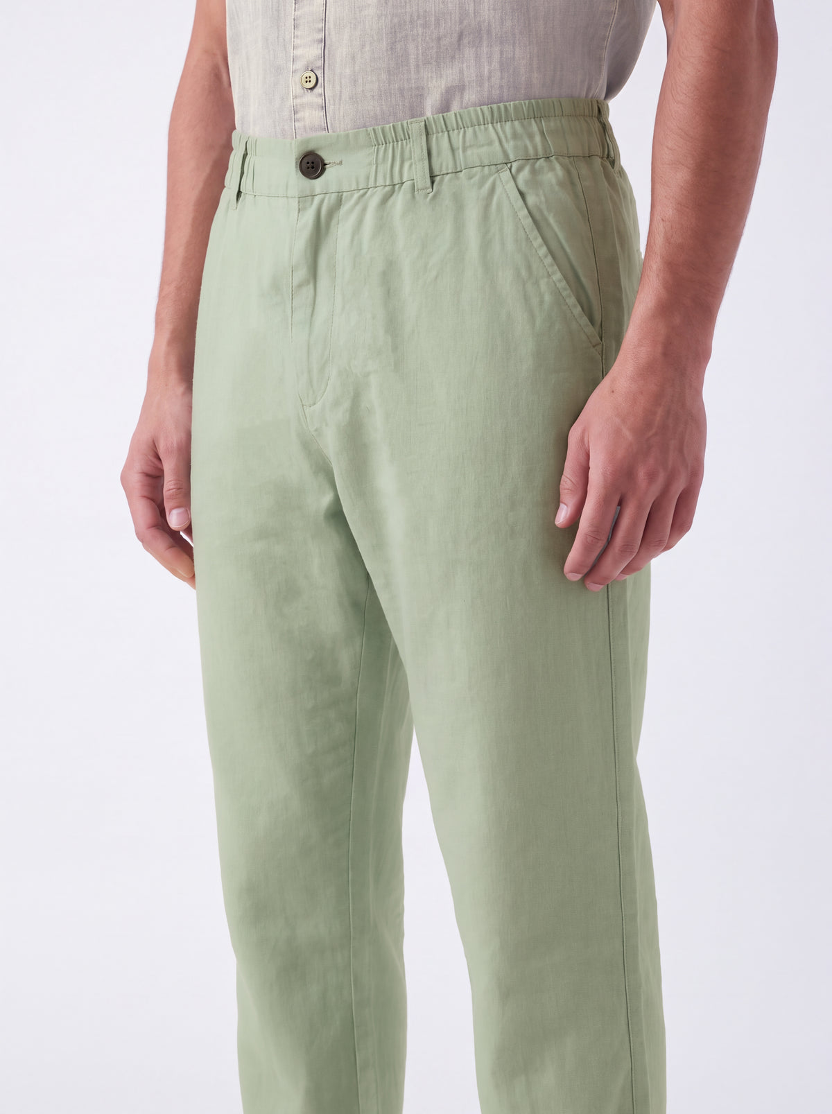 A person wearing light sage green trousers and a light beige button-up shirt, shown from the waist down against a plain white studio background.