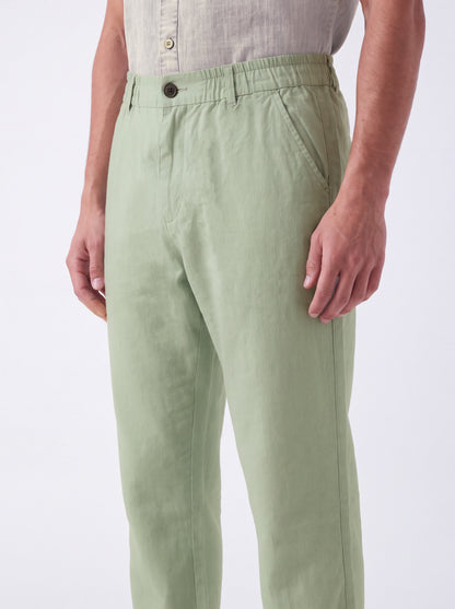 A person wearing light sage green trousers and a light beige button-up shirt, shown from the waist down against a plain white studio background.