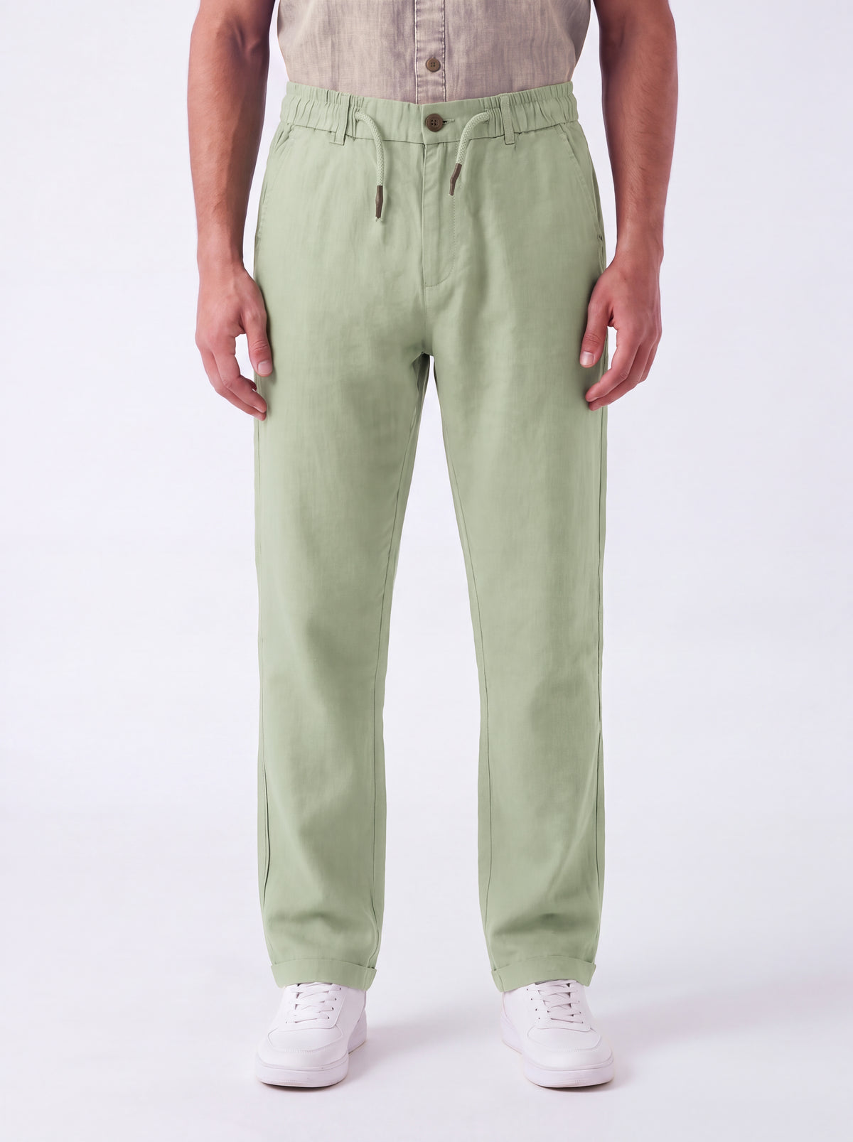A person wearing light sage green drawstring trousers, a beige shirt, and white sneakers, standing against a plain white studio background.