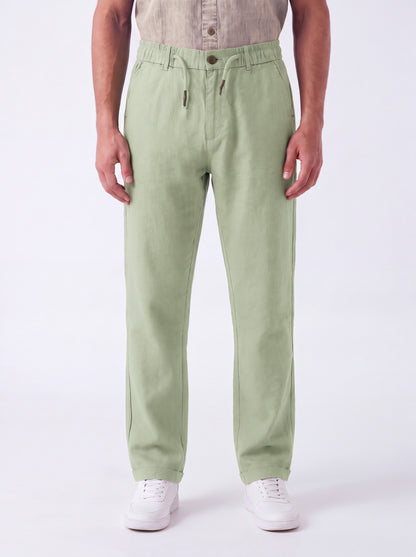 A person wearing light sage green drawstring trousers, a beige shirt, and white sneakers, standing against a plain white studio background.