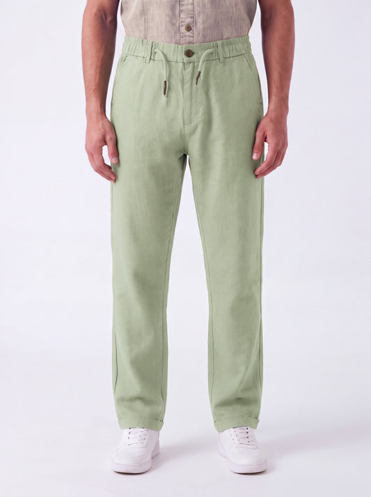 A person wearing light sage green drawstring trousers, a beige shirt, and white sneakers, standing against a plain white studio background.