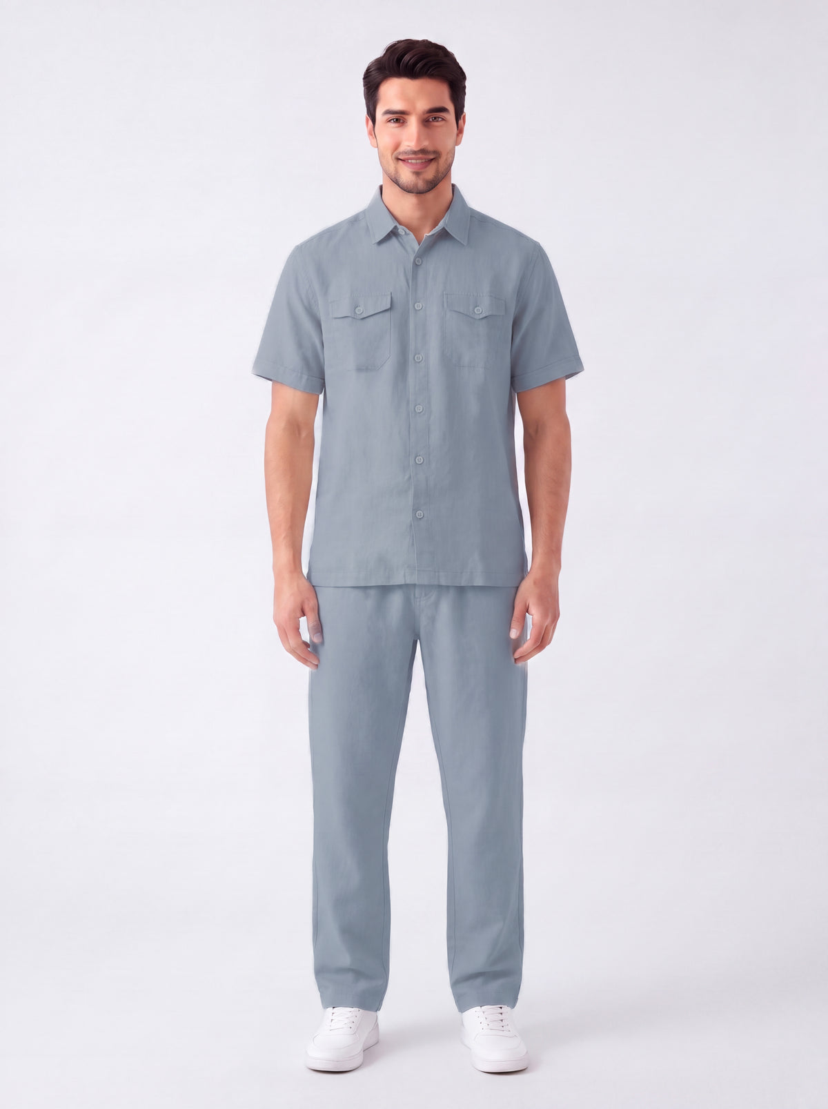 A man modeling a matching light blue short-sleeve button-up shirt and trousers set, paired with white sneakers, against a plain light gray studio background.