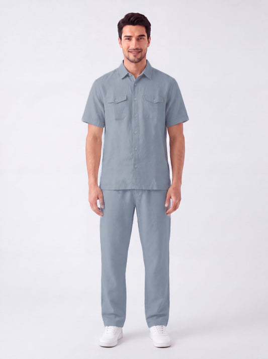 A man modeling a matching light blue short-sleeve button-up shirt and trousers set, paired with white sneakers, against a plain light gray studio background.