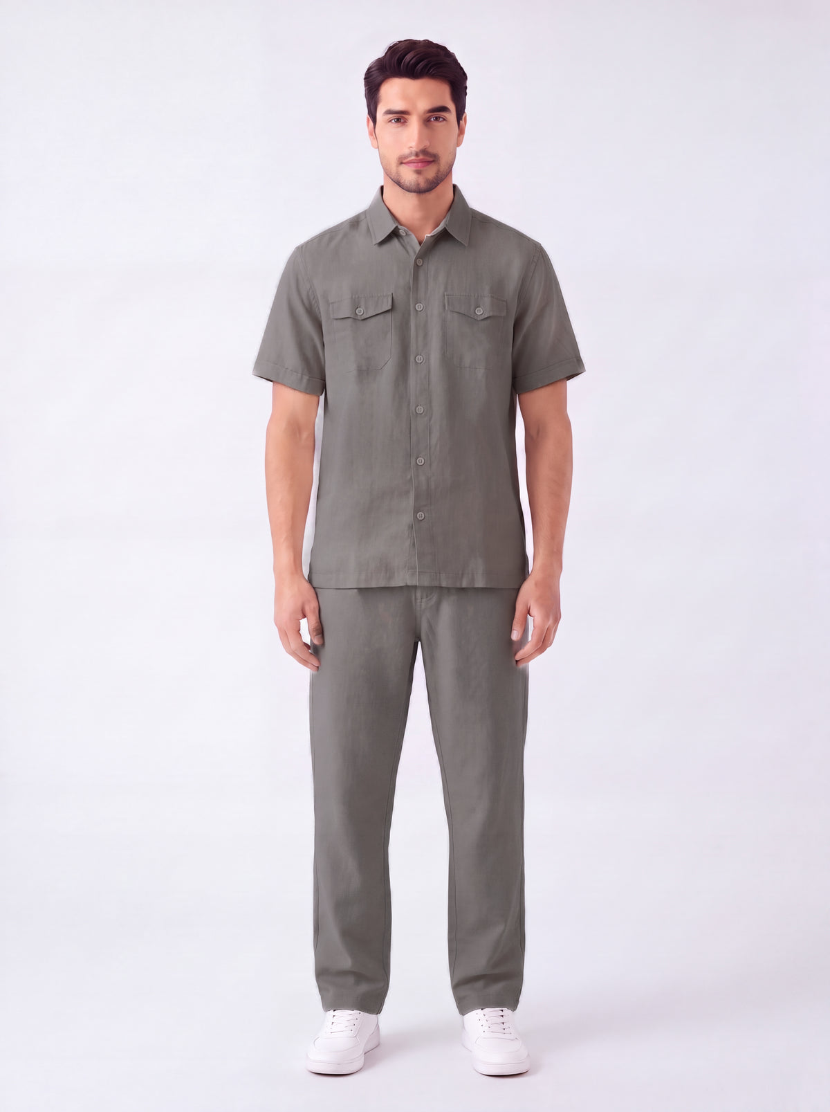 A man modeling a matching short-sleeve button-up shirt and trousers set in a solid olive green color, paired with white sneakers, against a plain white studio background.