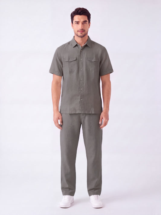 A man modeling a matching short-sleeve button-up shirt and trousers set in a solid olive green color, paired with white sneakers, against a plain white studio background.