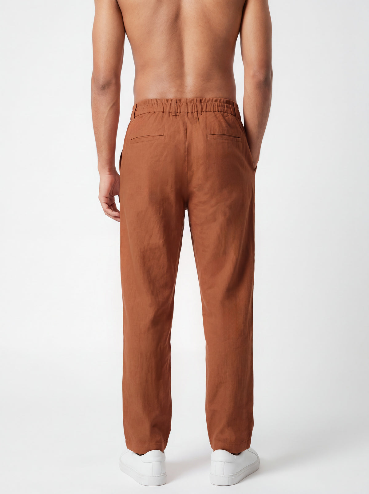 Rear view of a man wearing rust-brown linen trousers and white sneakers against a clean white studio background.