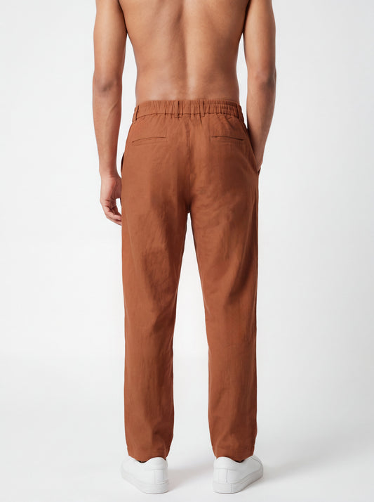 Rear view of a man wearing rust-brown linen trousers and white sneakers against a clean white studio background.