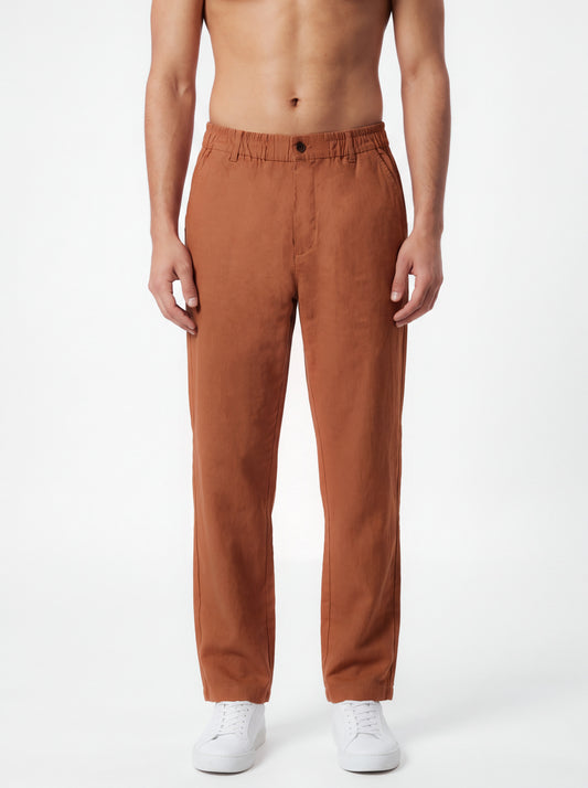 A man wearing rust-colored straight-leg trousers and white sneakers, standing against a plain white studio background.