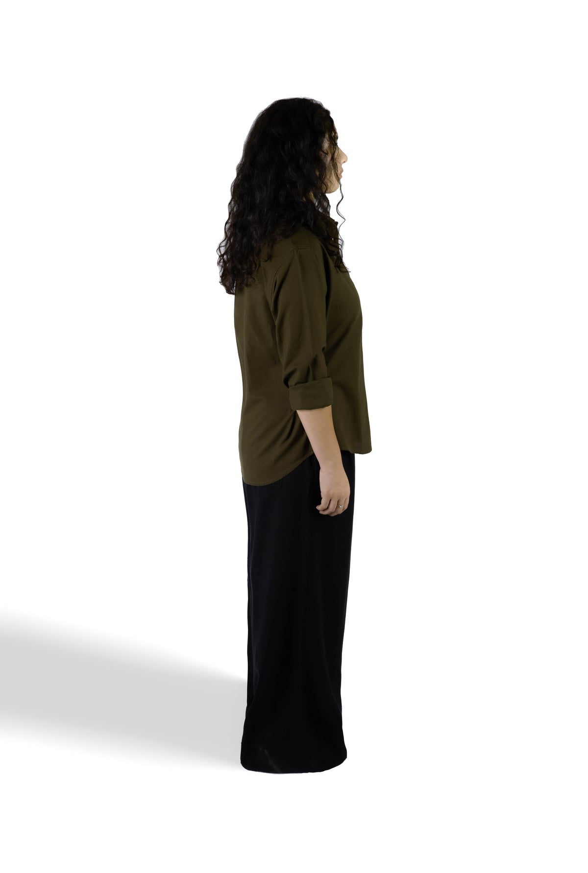 A woman shown from the side profile wearing a dark olive green long-sleeve top with rolled sleeves and a long black skirt, standing against a plain white studio background.