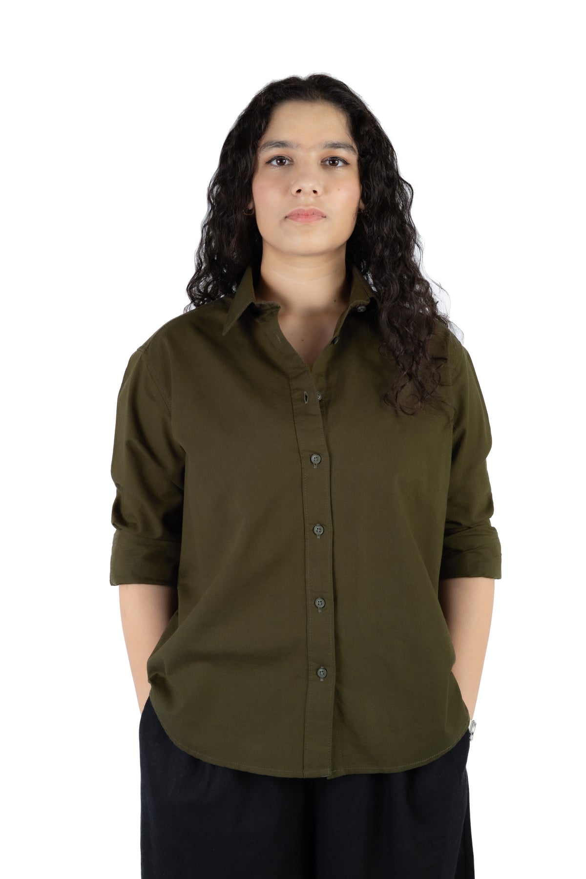 A woman with long, curly dark hair wearing an olive green button-down shirt with rolled-up sleeves and black pants, standing against a solid white background.