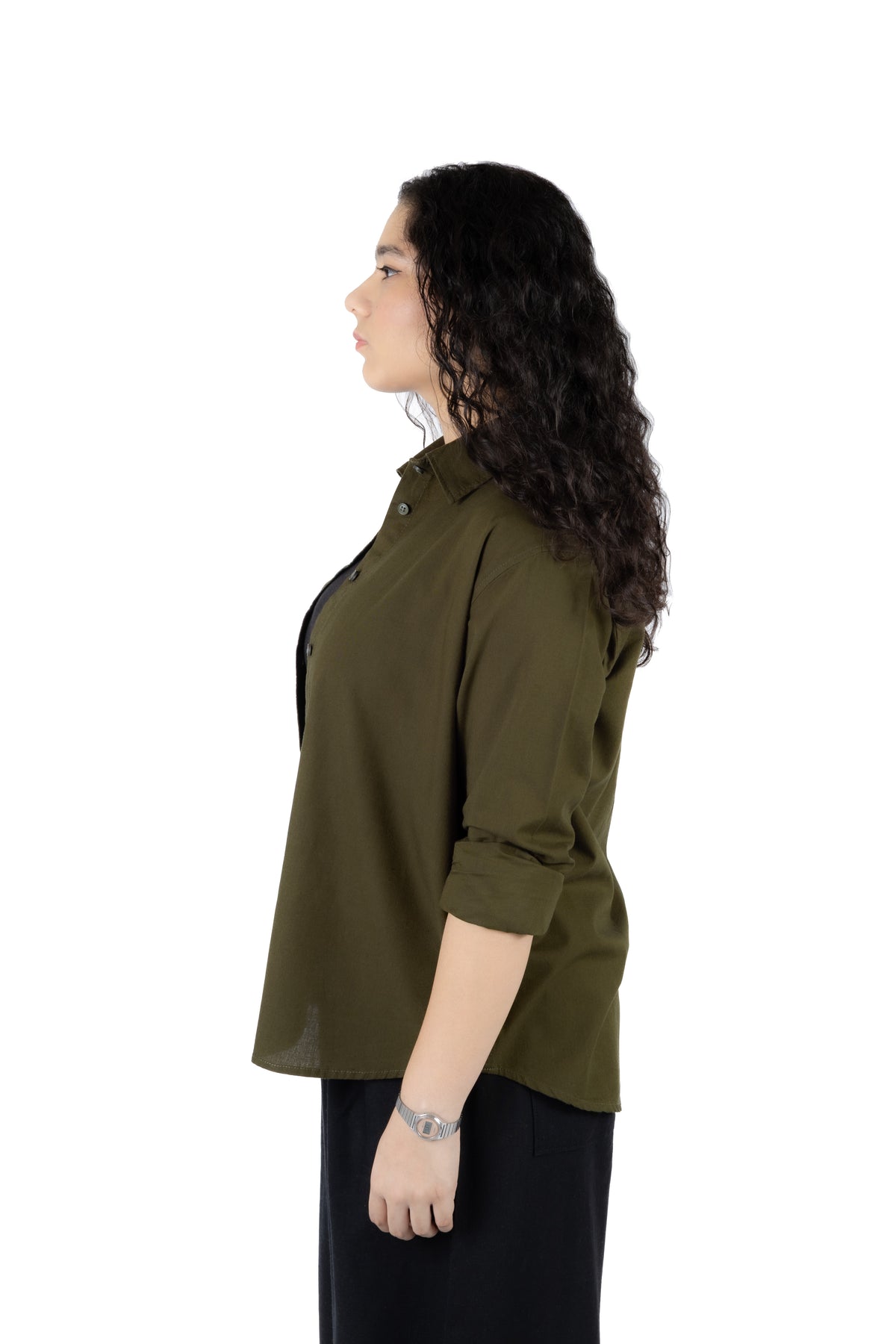 A woman with long, curly dark hair is shown in profile wearing an olive green button-up shirt with three-quarter sleeves and black pants, photographed against a solid white background.