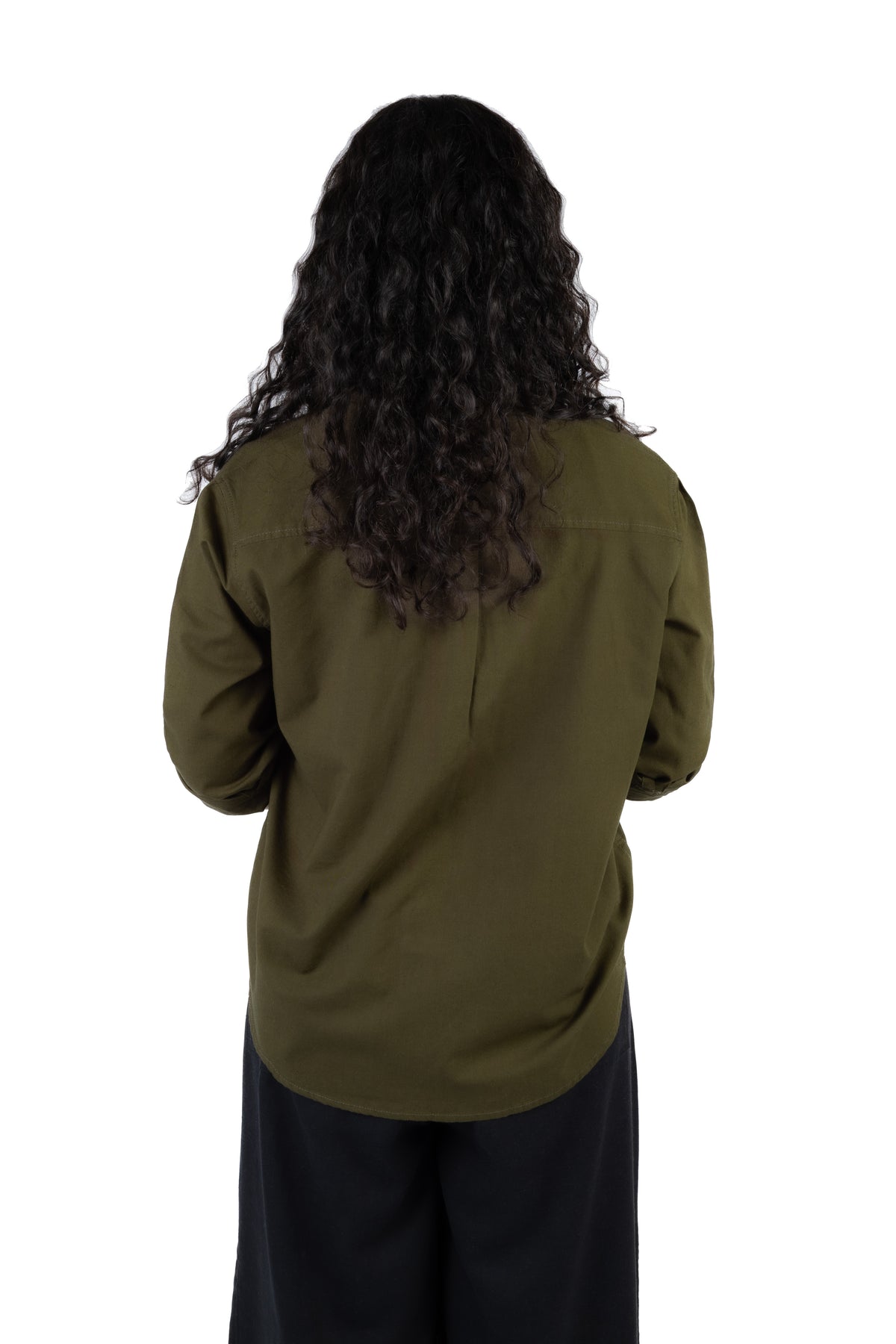 A person viewed from the back wearing a dark olive green long-sleeve shirt and black pants, with long dark curly hair, against a plain white background.