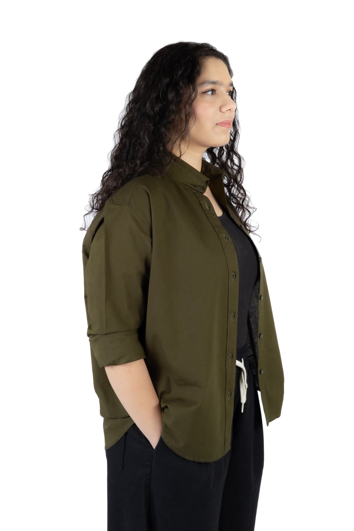 A woman with long, curly dark hair is shown in profile wearing an olive green button-up shirt over a black top and black pants, set against a plain white background.