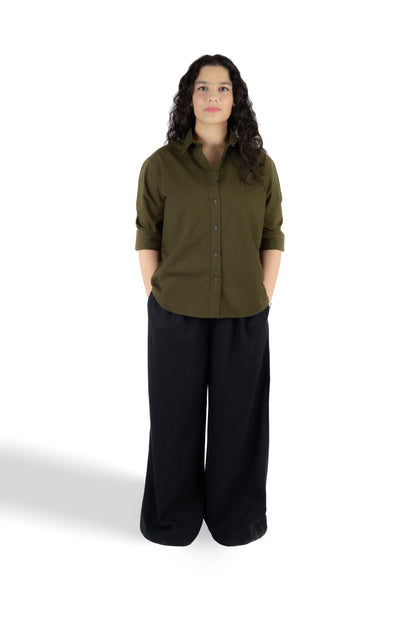 A woman stands against a white studio background wearing an olive green long-sleeve button-up shirt with rolled sleeves and black wide-leg pants.