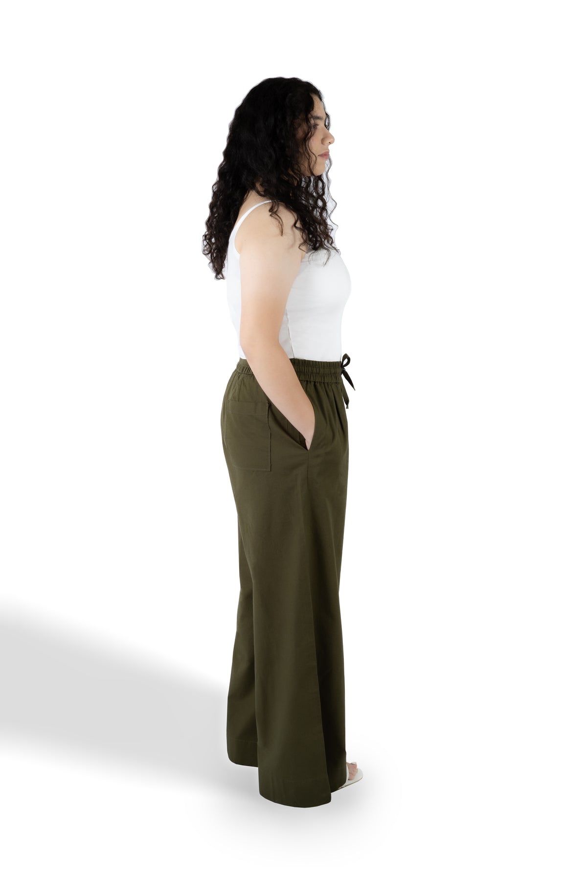 A woman shown from the side profile wearing olive green wide-leg pants with a drawstring waist and a white tank top, standing against a plain white studio background.