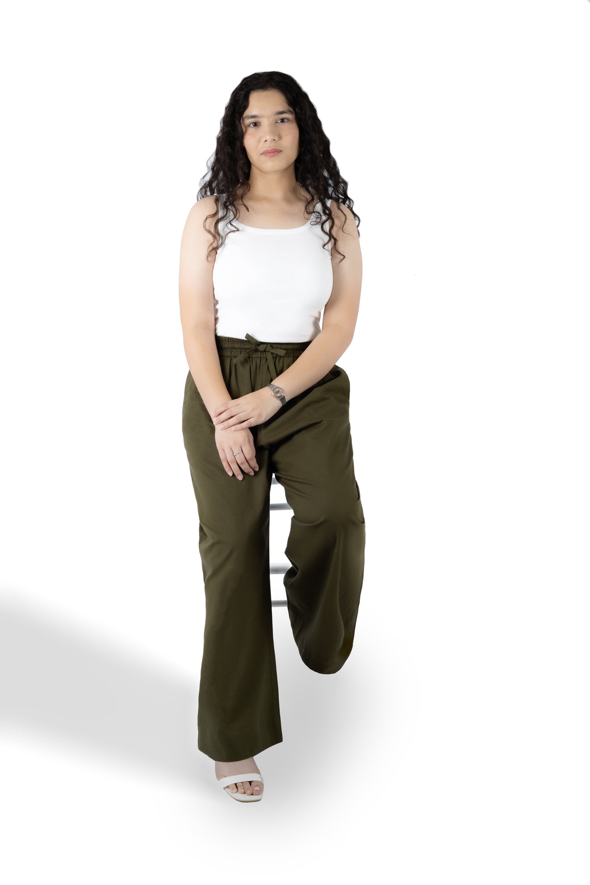A woman with long curly dark hair stands on a white studio background wearing olive green wide-leg pants, a white sleeveless top, and white heeled sandals.