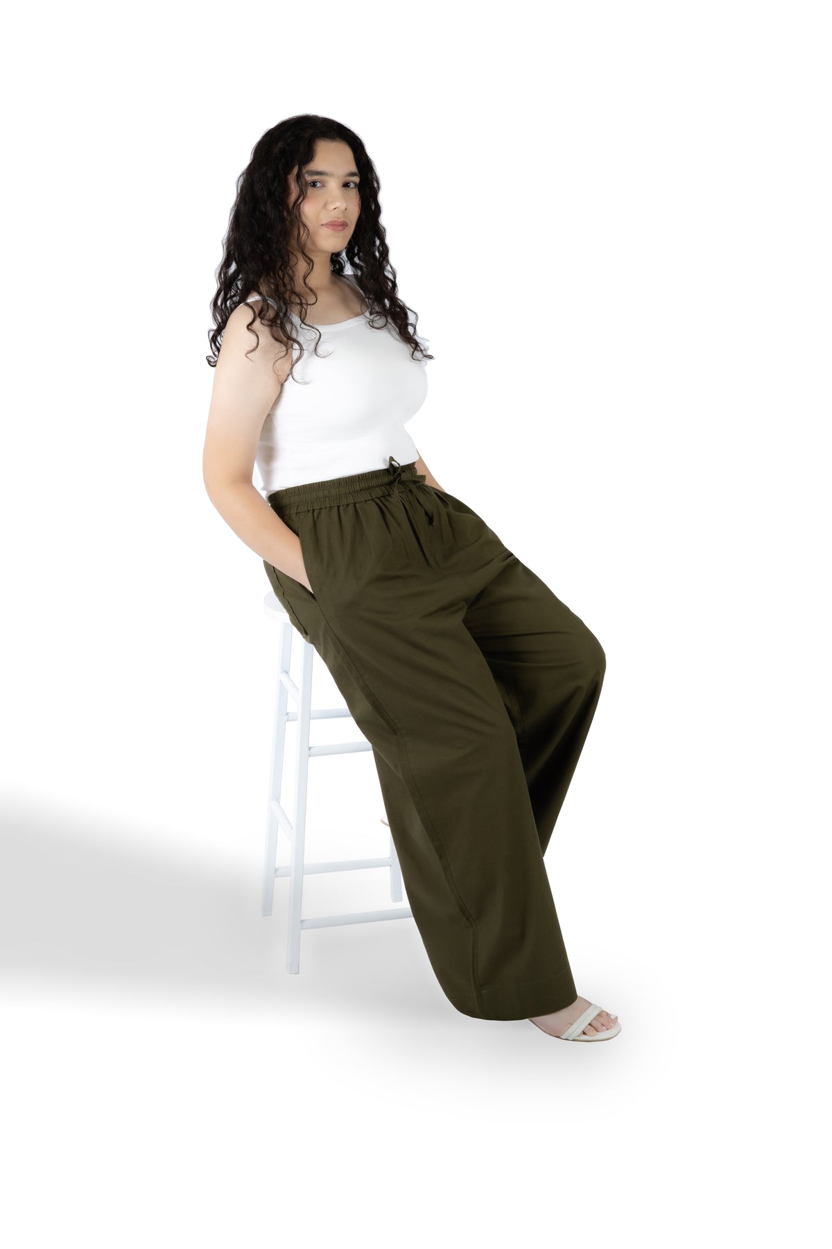 A woman with long curly dark hair sits on a white stool against a white studio background, wearing a white sleeveless top and olive green wide-leg drawstring pants.