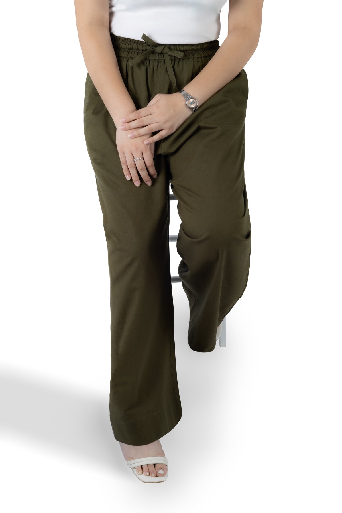 A person wearing olive green wide-leg drawstring pants, styled with a white top and white heeled sandals, seated on a white stool against a white background.