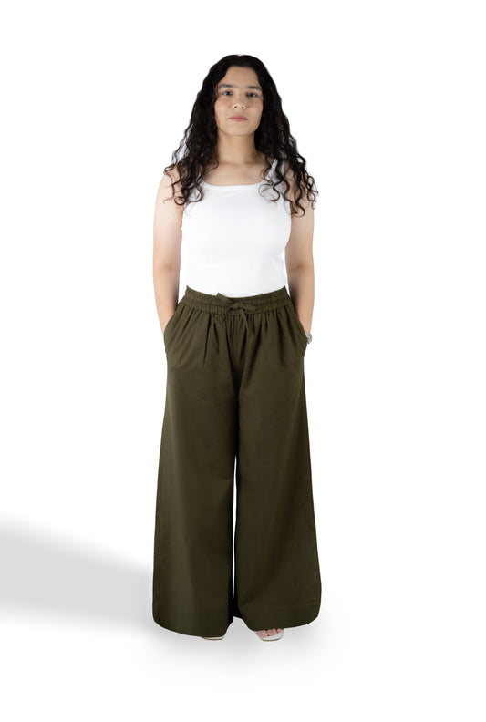 A woman modeling a pair of wide-leg olive green pants with a white sleeveless top, standing against a plain white studio background.