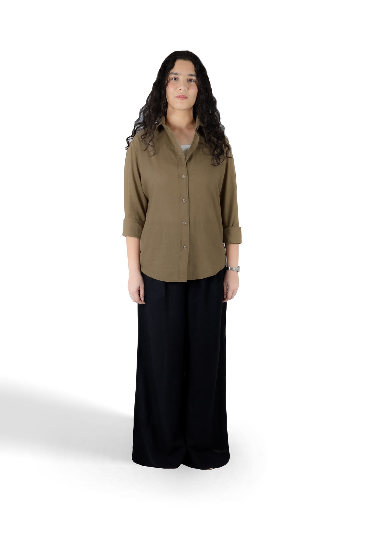 A woman stands against a plain white studio background wearing a muted olive-brown button-up shirt with three-quarter sleeves and black wide-leg pants.