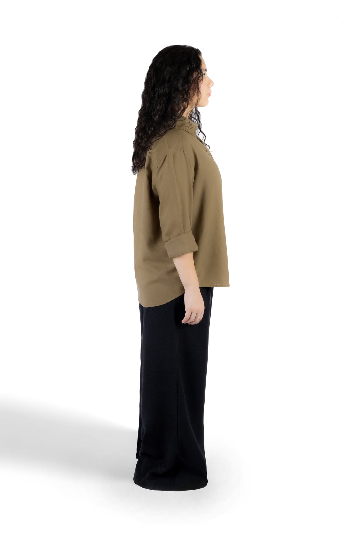 A woman shown in side profile wearing a loose olive-brown long-sleeve top with rolled sleeves and black wide-leg pants, standing against a plain white studio background.