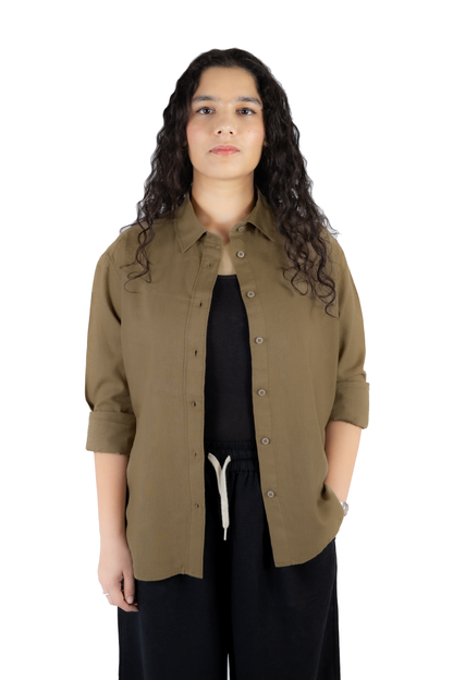 A woman with long, curly dark hair stands against a solid white background wearing an olive green button-up shirt over a black top and black pants with a white drawstring.