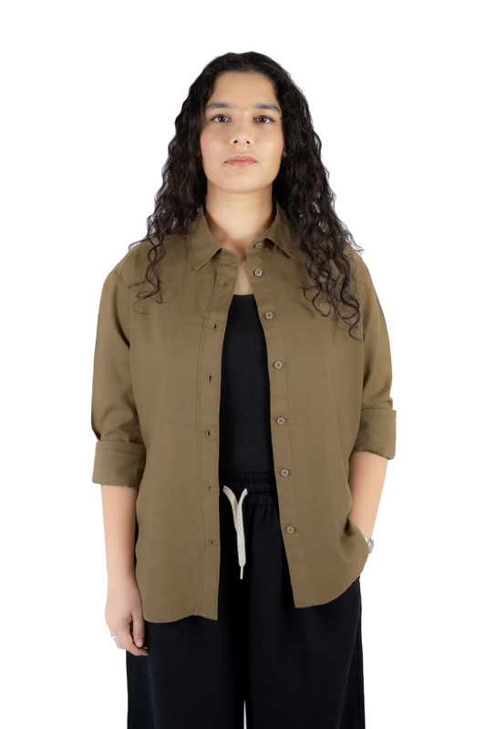 A woman with long, curly dark hair stands against a solid white background wearing an olive green button-up shirt over a black top and black pants with a white drawstring.