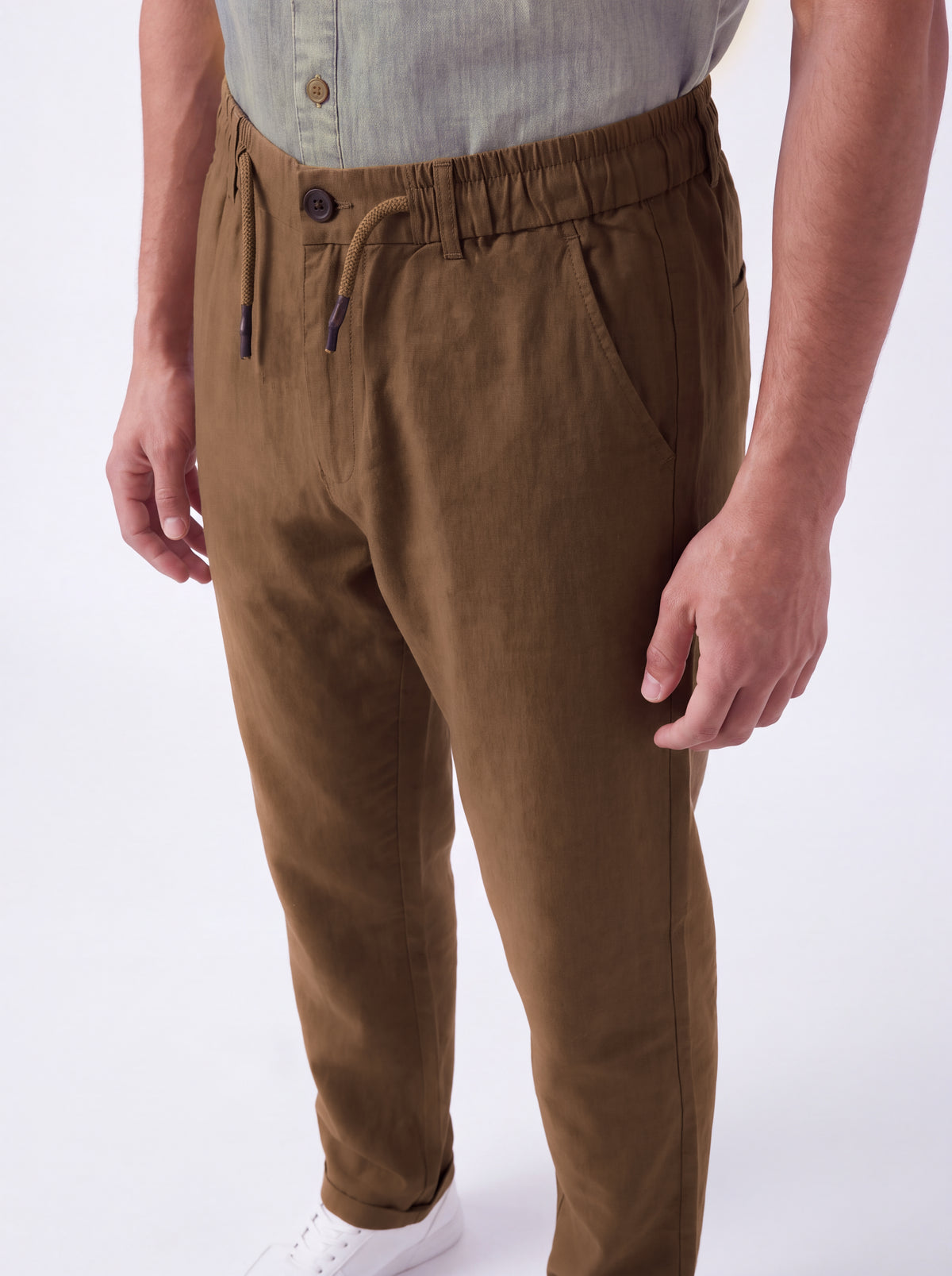 A person wearing brown drawstring trousers with a relaxed fit, paired with a light gray shirt and white sneakers, shown from the waist down against a plain white background.