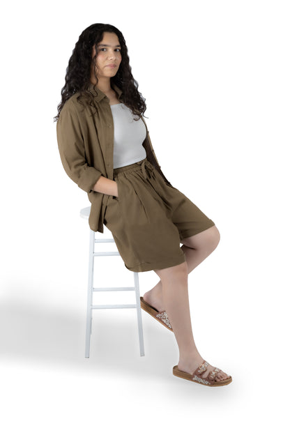 A woman with long curly dark hair sits on a white stool against a white studio background, wearing a matching brown linen-look short jacket and shorts set over a white top and brown slide sandals.