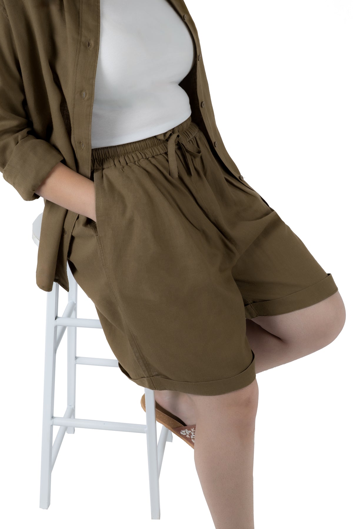 A person seated on a white stool wearing olive brown drawstring shorts with a white top and an open olive brown shirt, photographed against a plain white background.