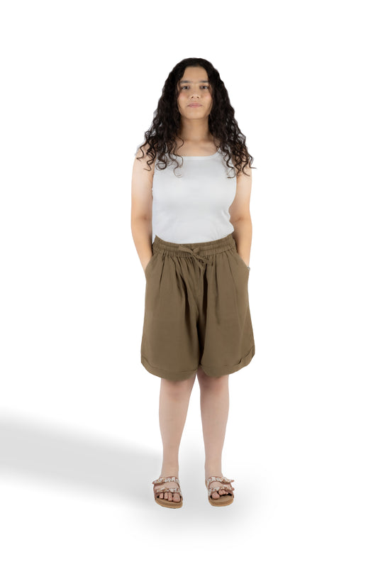 A woman with long curly dark hair stands against a white studio background wearing a white sleeveless top, brown drawstring shorts, and brown sandals.