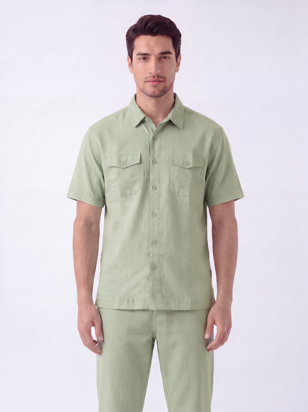 A man modeling a light sage green short-sleeve button-up shirt with two chest pockets, paired with matching light green trousers, against a plain white studio background.