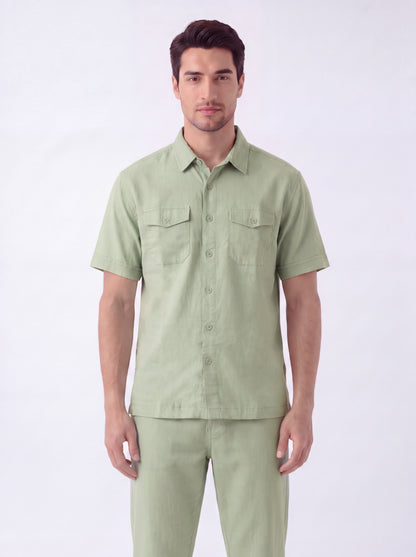 A man modeling a light sage green short-sleeve button-up shirt with two chest pockets, paired with matching light green trousers, against a plain white studio background.