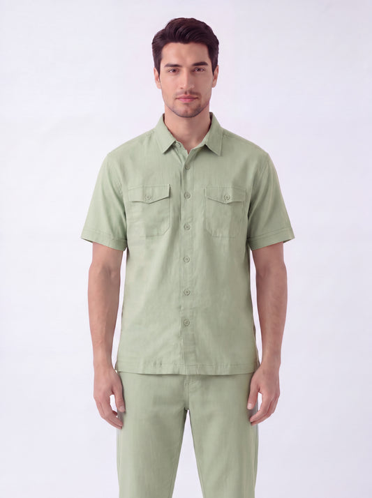 A man modeling a light sage green short-sleeve button-up shirt with two chest pockets, paired with matching light green trousers, against a plain white studio background.