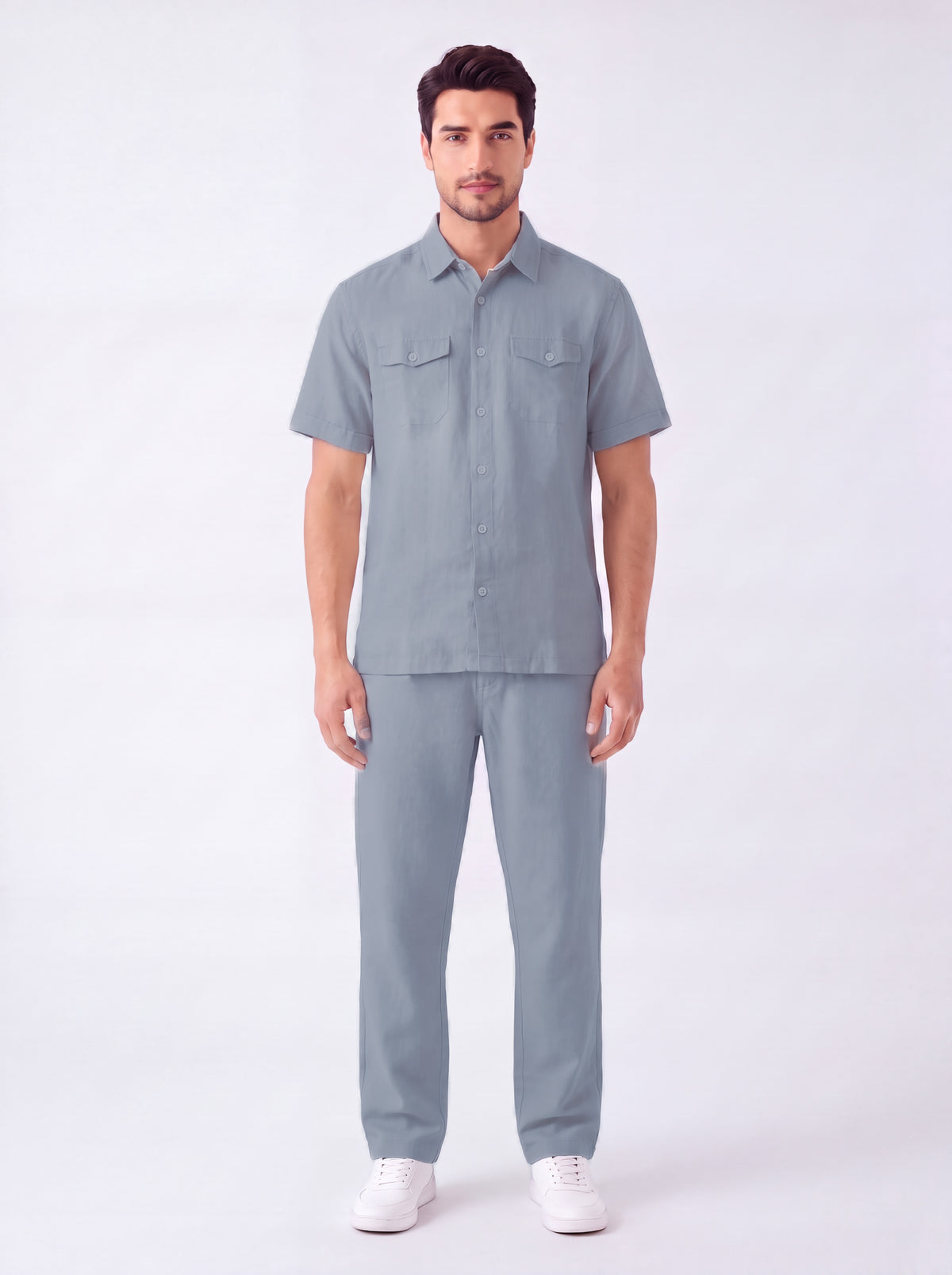 A man modeling a matching light blue-gray short-sleeve button-up shirt and trousers set, paired with white sneakers, against a plain white studio background.