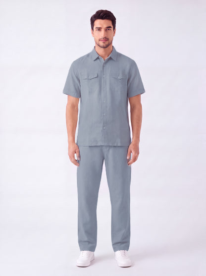 A man modeling a matching light blue-gray short-sleeve button-up shirt and trousers set, paired with white sneakers, against a plain white studio background.