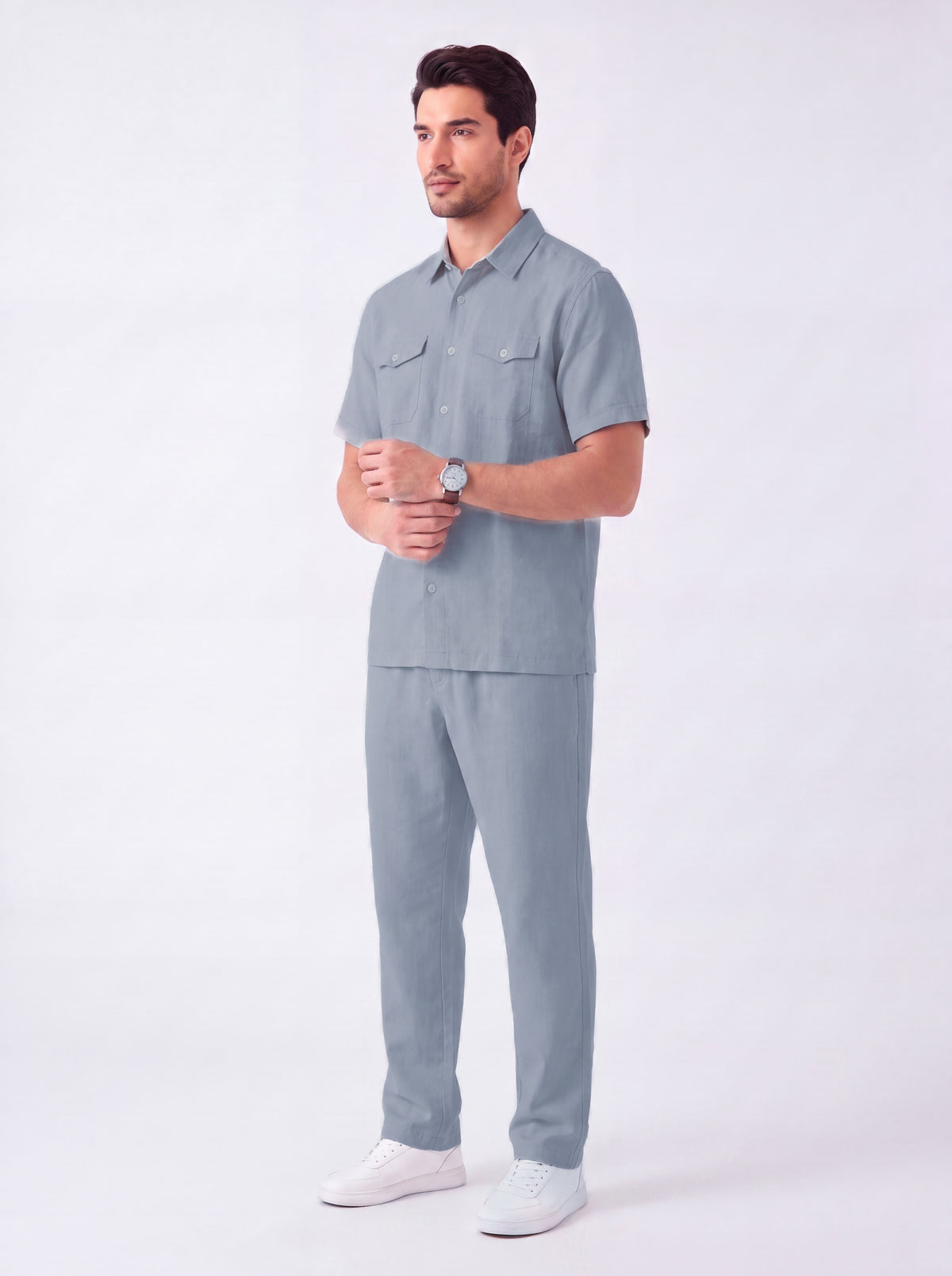 A man modeling a light blue-gray short-sleeve button-up shirt and matching pants set, paired with white sneakers, against a plain light gray studio background.