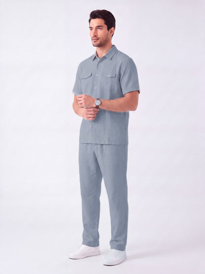 A man modeling a light blue-gray short-sleeve button-up shirt and matching pants set, paired with white sneakers, against a plain light gray studio background.
