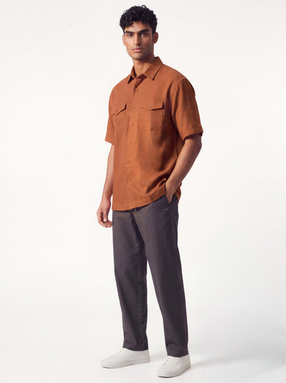 A man modeling a short-sleeve button-up shirt in rust orange paired with dark gray trousers and white sneakers, standing against a clean white studio background.