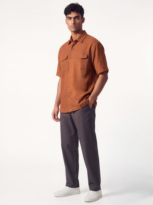 A man modeling a short-sleeve button-up shirt in rust orange paired with dark gray trousers and white sneakers, standing against a clean white studio background.